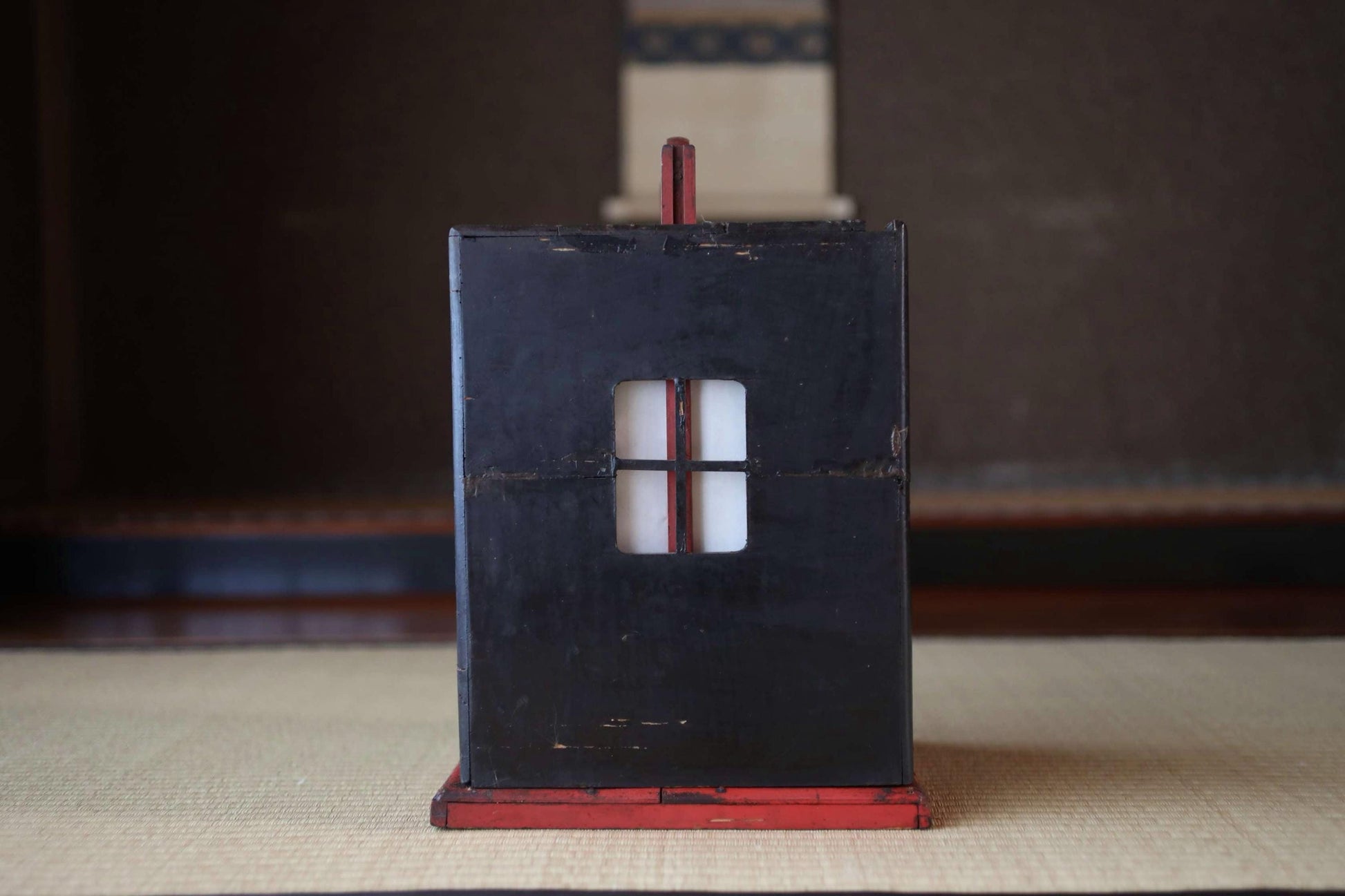 A black wooden box with a red handle, featuring a small window, sits on a beige surface.