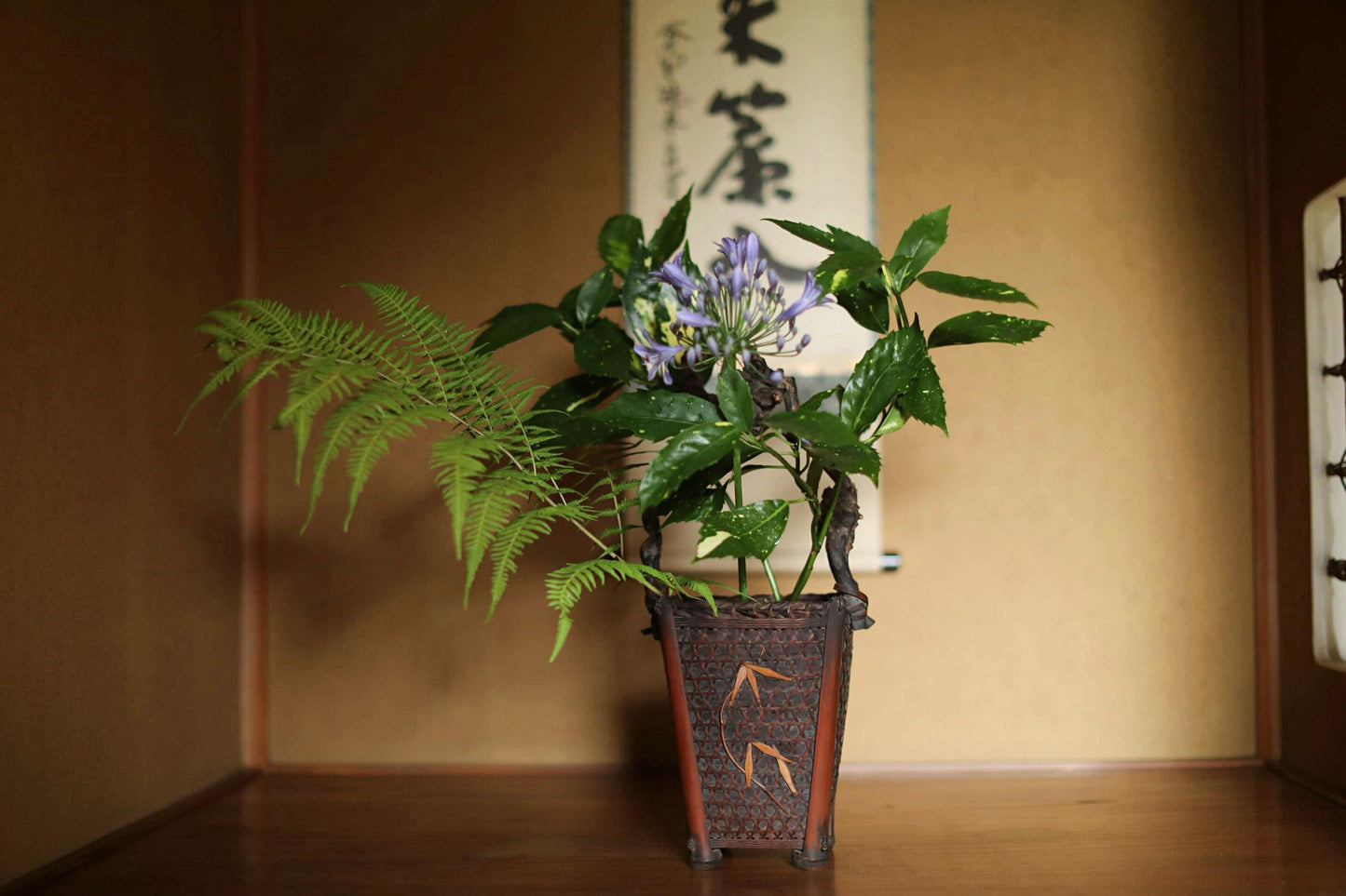 A potted plant with green leaves and purple flowers sits on a wooden surface, with a traditional Japanese-style banner hanging in the background.