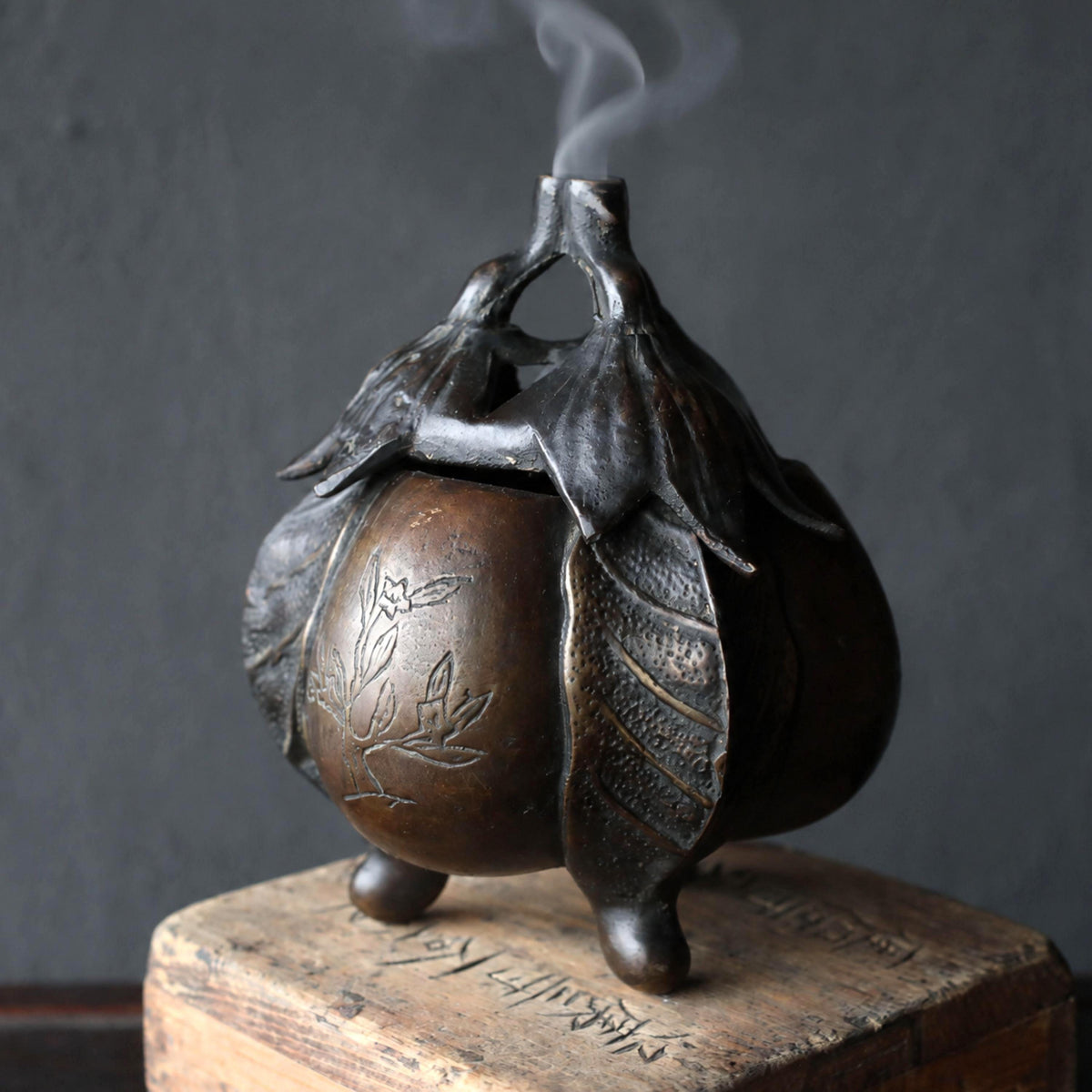 A bronze incense burner with a floral design sits on a wooden surface, emitting smoke.