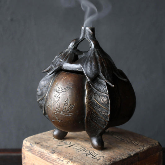 A bronze incense burner with a floral design sits on a wooden surface, emitting smoke.