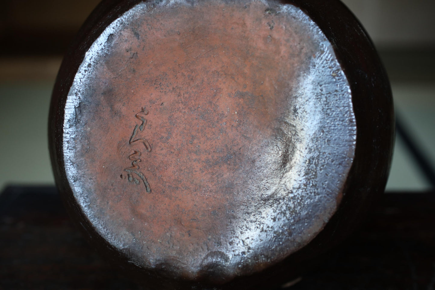the back of a dark-colored ceramic object, possibly a vase or bowl, with a textured surface and a small inscription or mark on it.