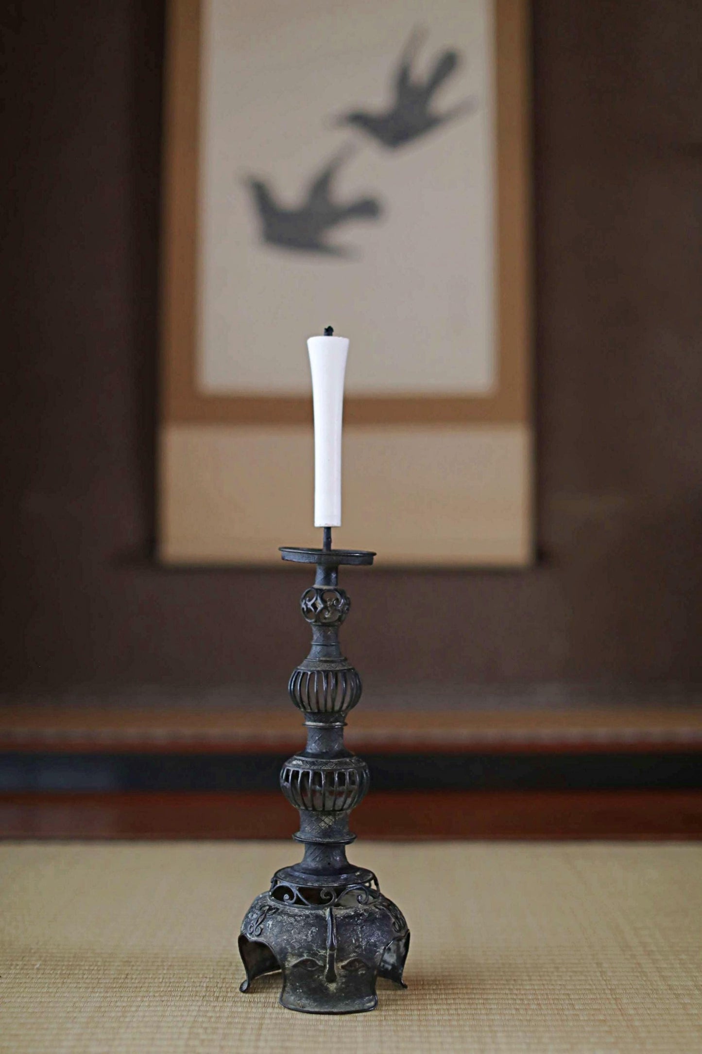 A white candle with a tall, ornate metal candlestick stands on a woven mat in the foreground, with a framed image of two birds in the background.