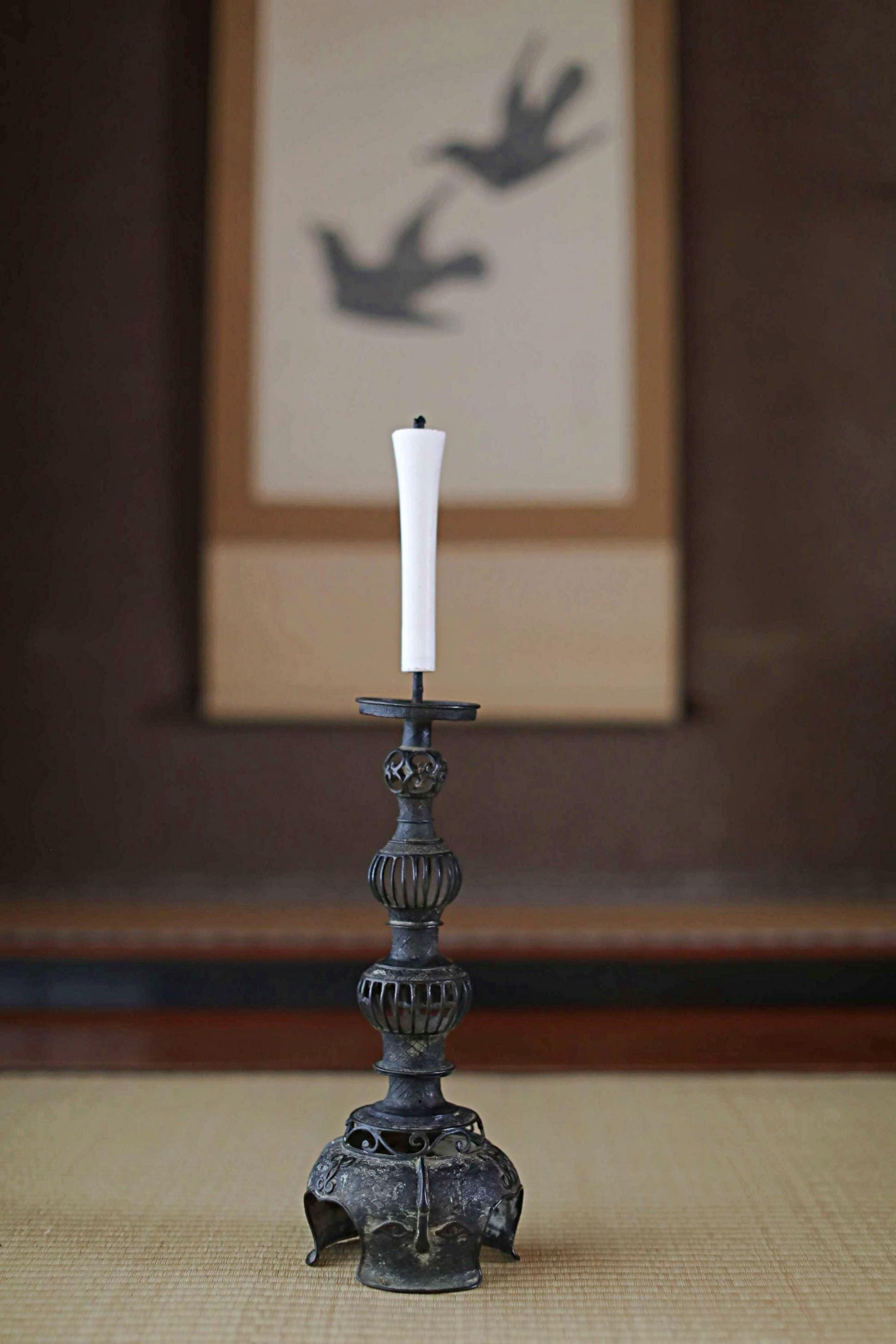 A white candle with a tall, ornate metal candlestick stands on a woven mat in the foreground, with a framed image of two birds in the background.