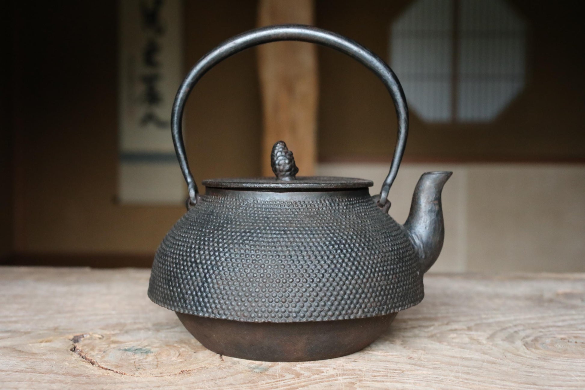 A black metal teapot with a textured surface sits on a wooden surface, with a blurred background suggesting an indoor setting.