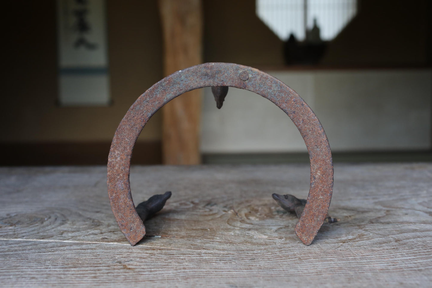 An old, rusted horseshoe sits on a wooden surface, with a blurred background that includes a window and a wall with Japanese characters.