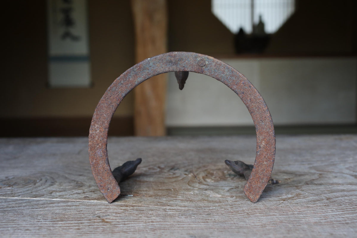 An old, rusted horseshoe sits on a wooden surface, with a blurred background that includes a window and a wall with Japanese characters.