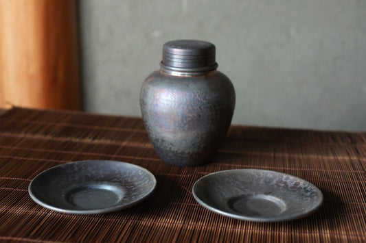 a dark-colored metal container, possibly a vase or jar, placed on a brown woven placemat. There are two matching dark-colored plates or saucers placed in front of the container, creating a simple and elegant arrangement.