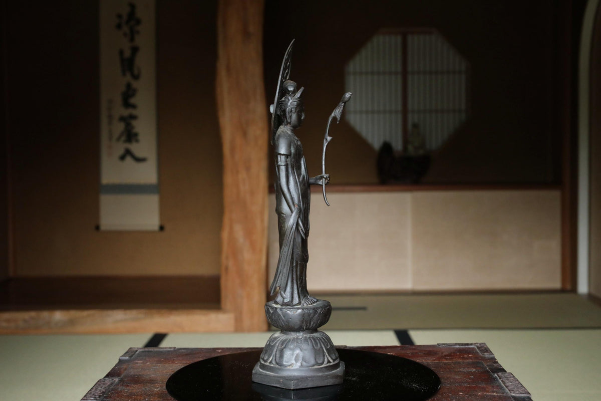 A bronze statue of a deity stands on a wooden base in a traditional Japanese temple setting.