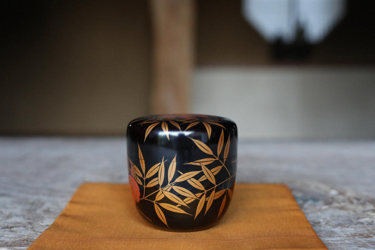A small, black ceramic container with a gold leaf design sits on a beige cloth.
