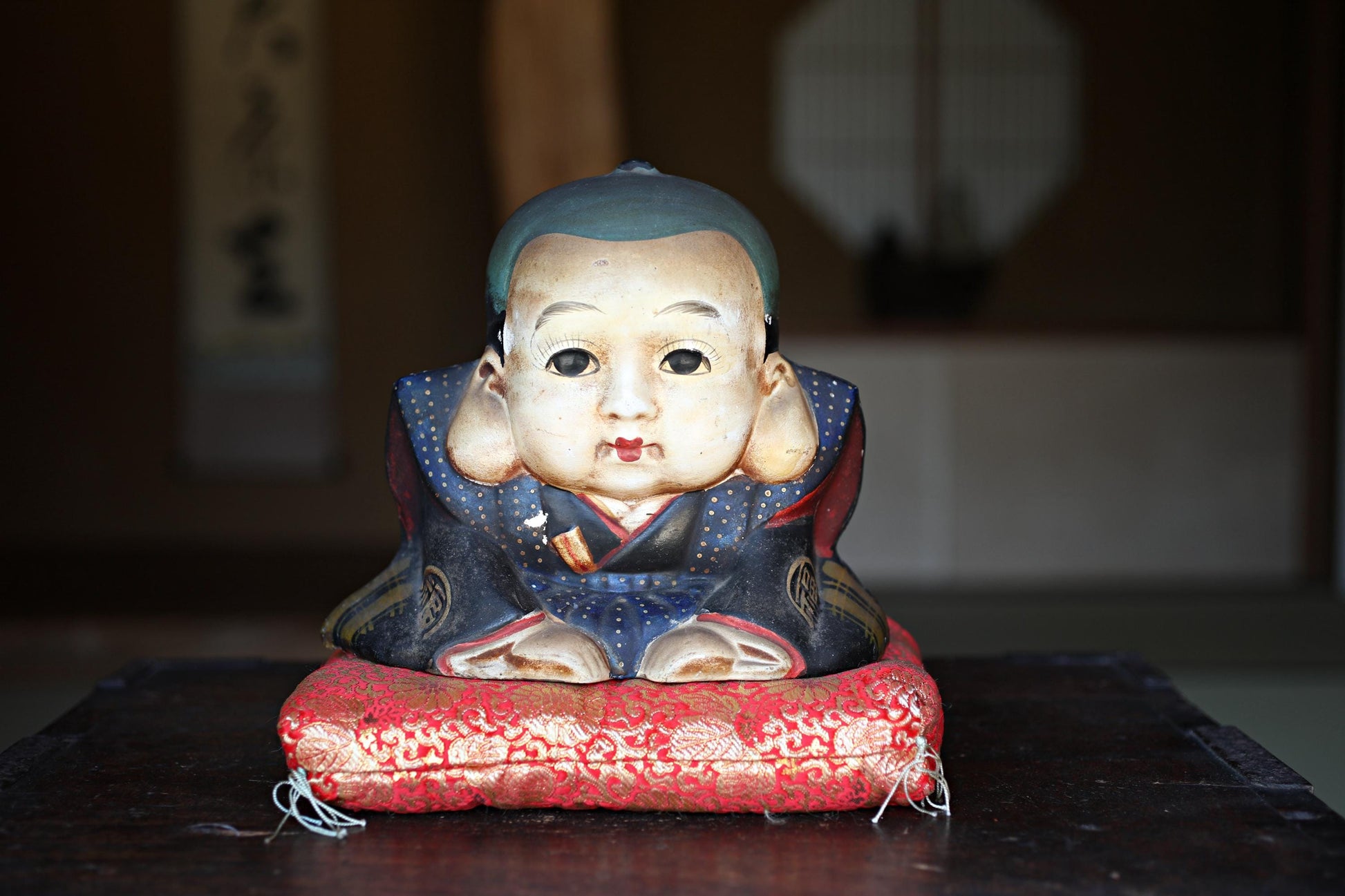 A ceramic figurine of a young boy sitting on a red cushion, placed on a wooden surface.