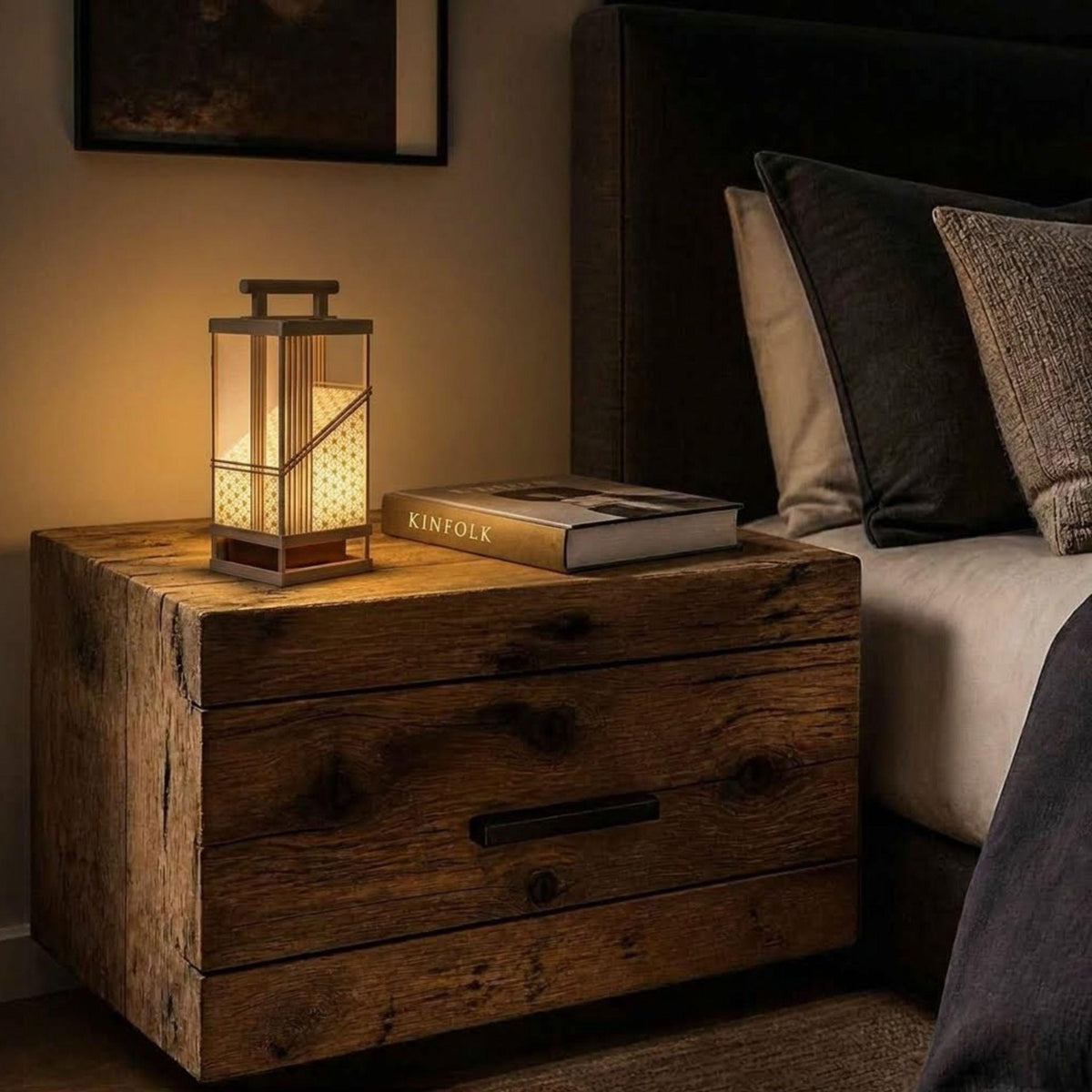 A wooden nightstand with a lit lantern, a book, and a pillow on it, situated next to a bed.