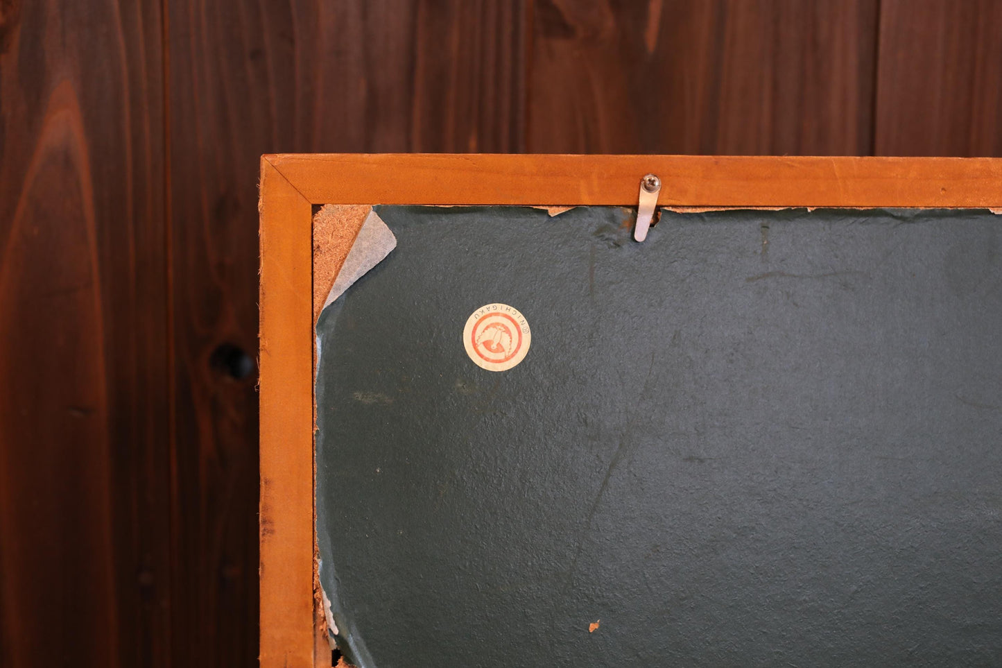 a close-up view of a wooden frame with a black background, and a small circular sticker or label is visible on the frame.