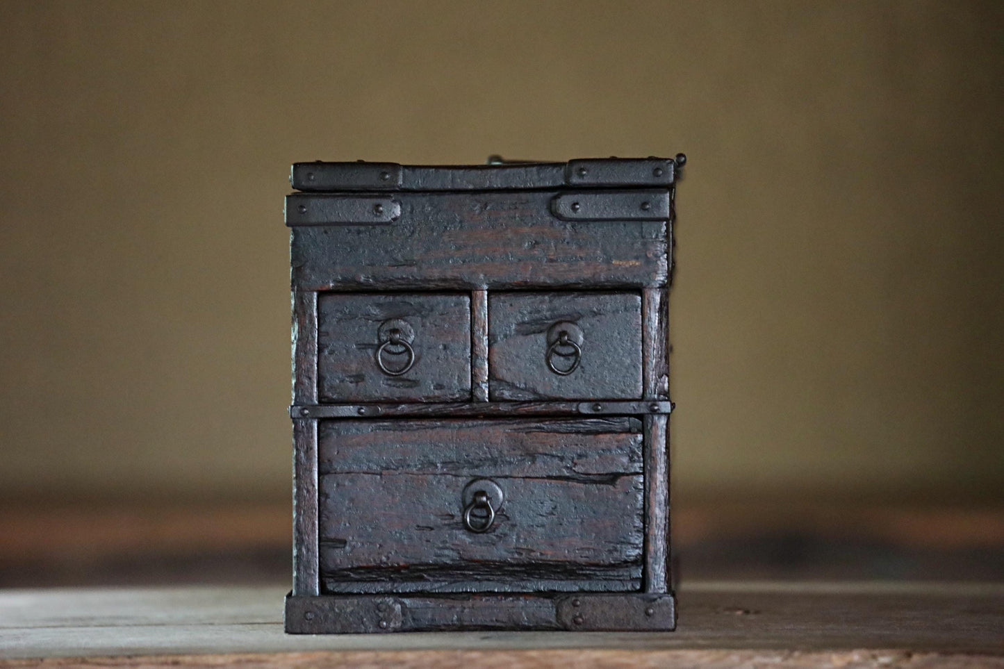 An old, weathered wooden chest with three drawers sits on a wooden surface against a blurred background.