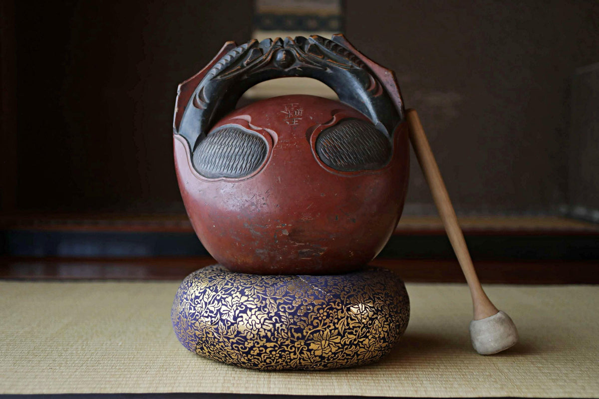 A red and black ceramic jug with a wooden handle, placed on a blue and gold decorative base, with a wooden stick leaning against it.