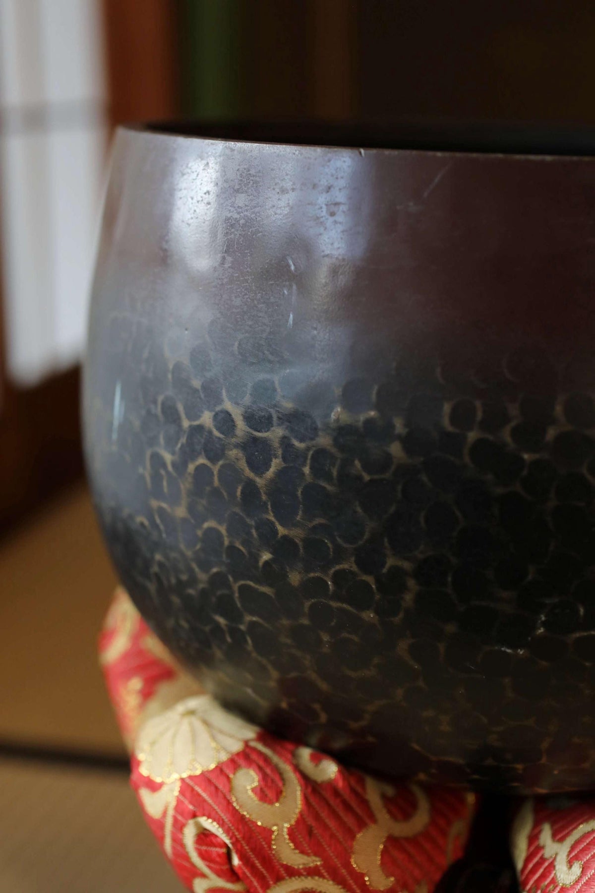 A close-up image of a dark-colored bowl or vase with a textured pattern, resting on a red and gold fabric surface.