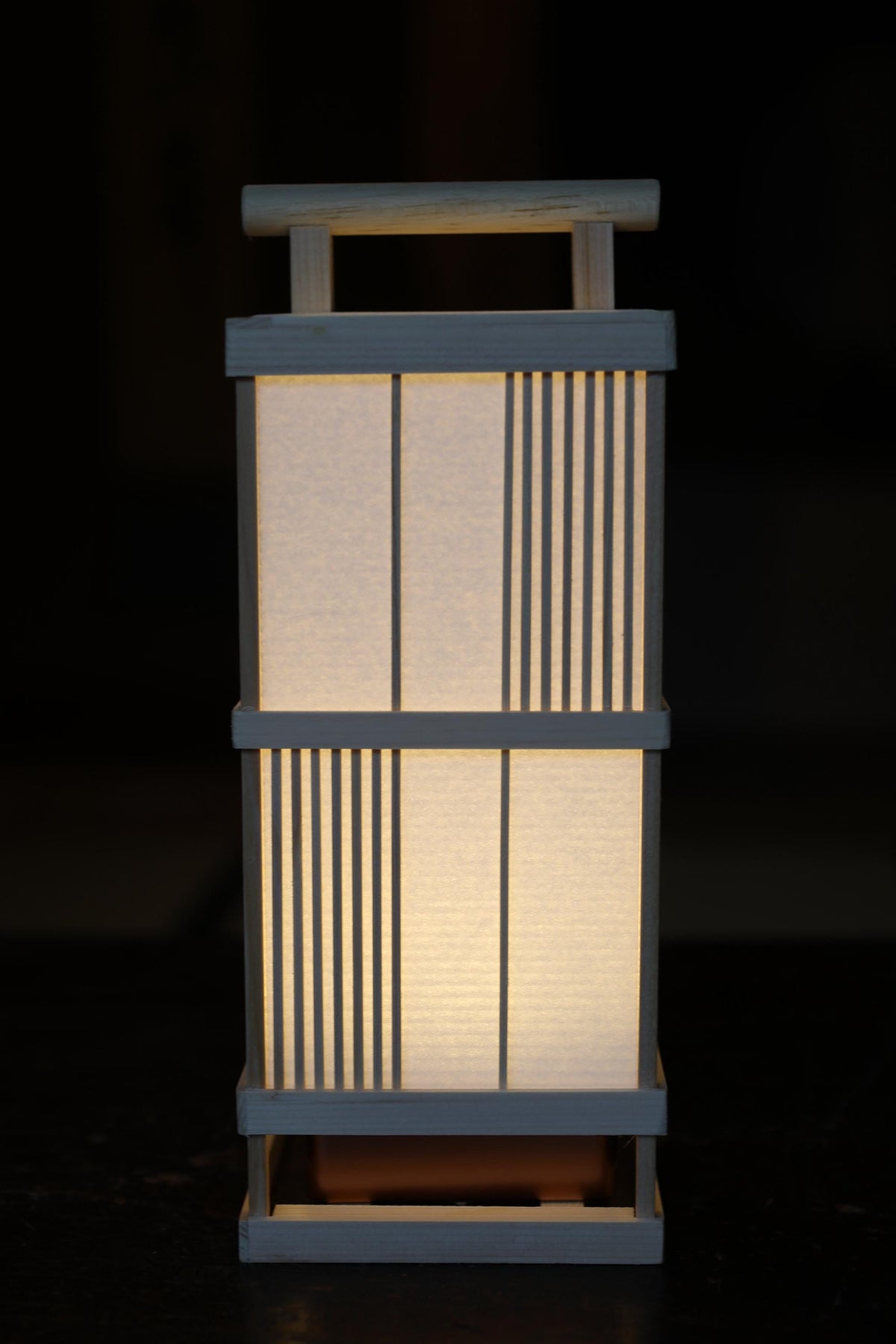 A white wooden lantern with a handle and light, standing alone against a dark background.