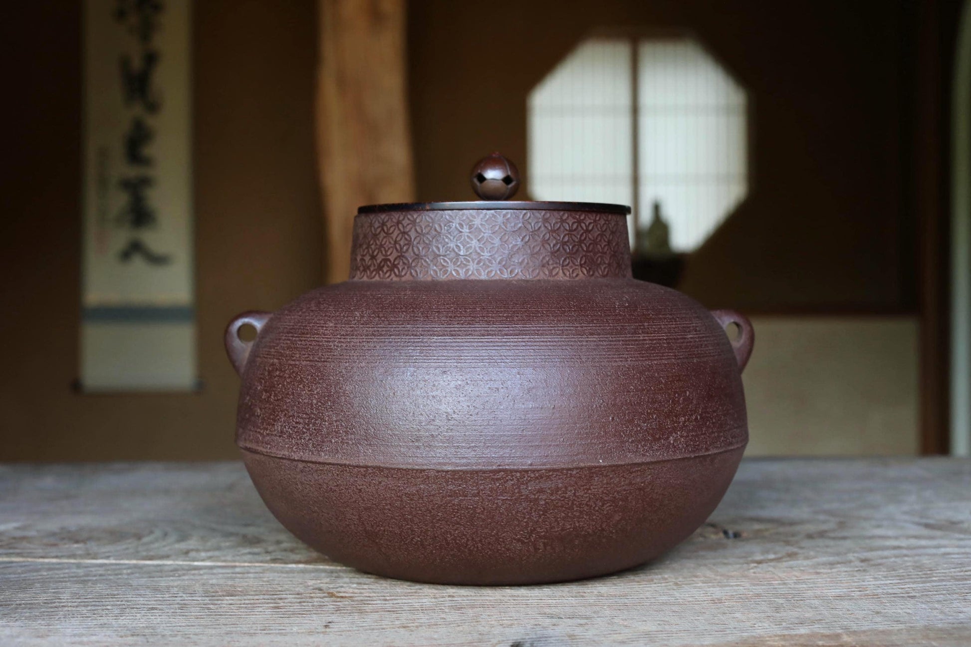 A large, round brown ceramic teapot with a lid sits on a wooden surface, with a window and a sign with Chinese characters visible in the background.