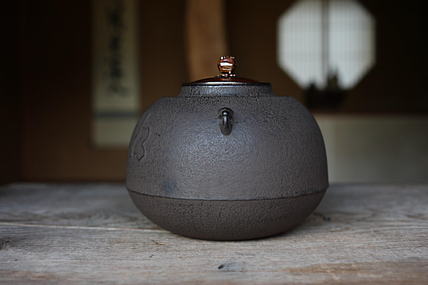 A black ceramic teapot with a metal lid sits on a wooden surface, with a blurred background that includes a window and a wall with Chinese characters.