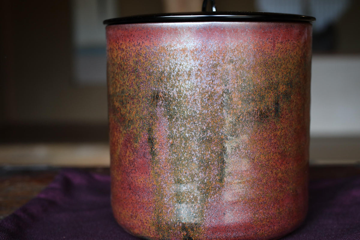A close-up image of a red and orange ceramic jar or container with a black lid, placed on a purple surface.