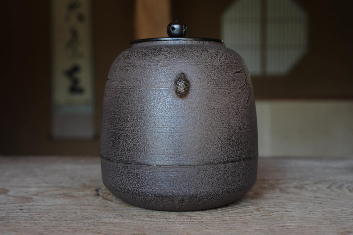 A ceramic teapot with a black lid sits on a wooden surface, with a blurred background that includes a window and a wall with Japanese characters.