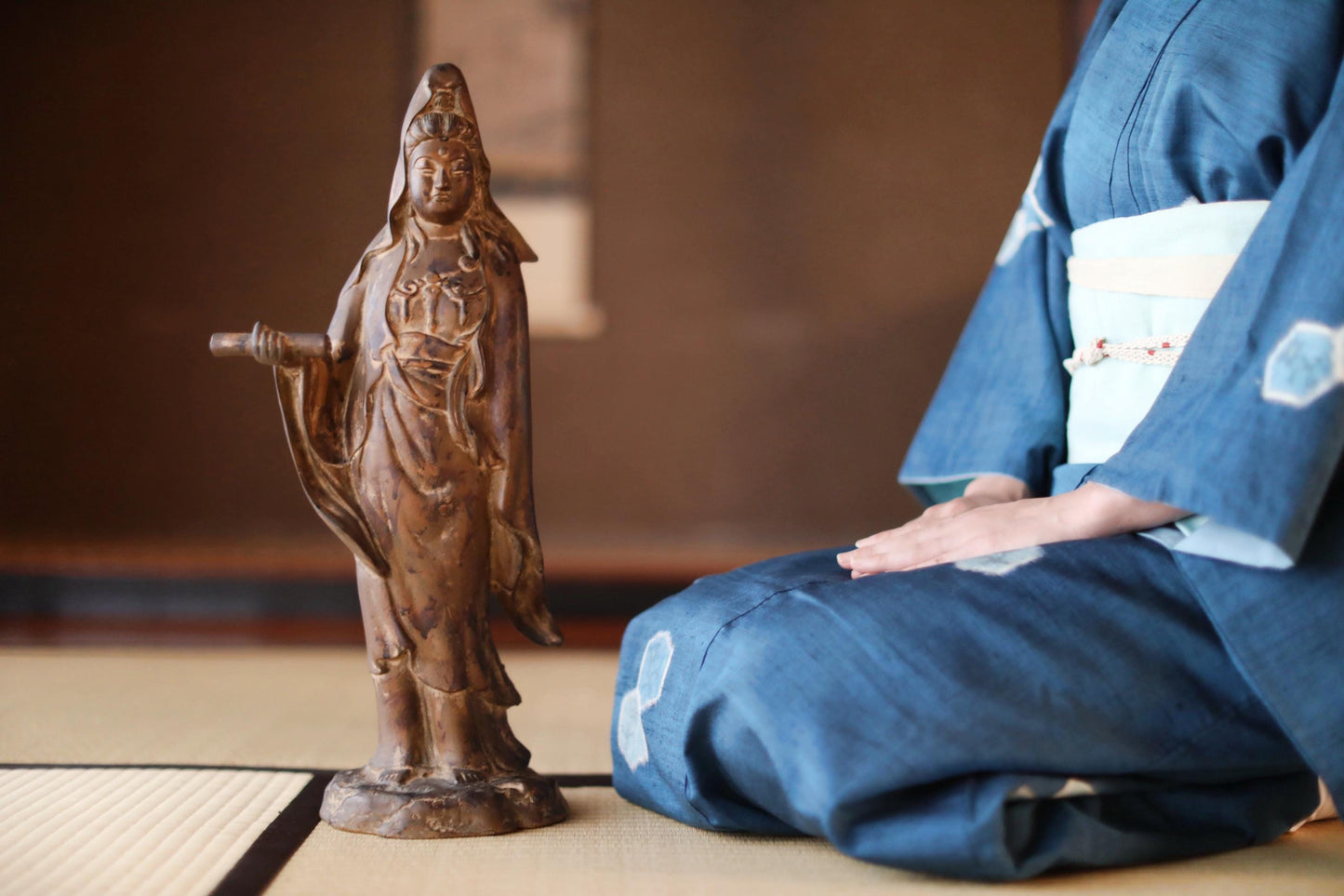 A person in a blue kimono is kneeling next to a small statue of a deity, which appears to be a wooden figurine.