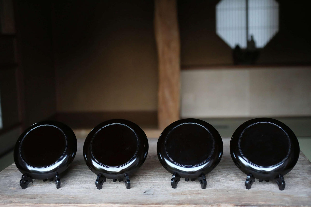 Four black ceramic plates or bowls are arranged in a row on a wooden surface, with a blurred background that includes a window and a wooden beam.