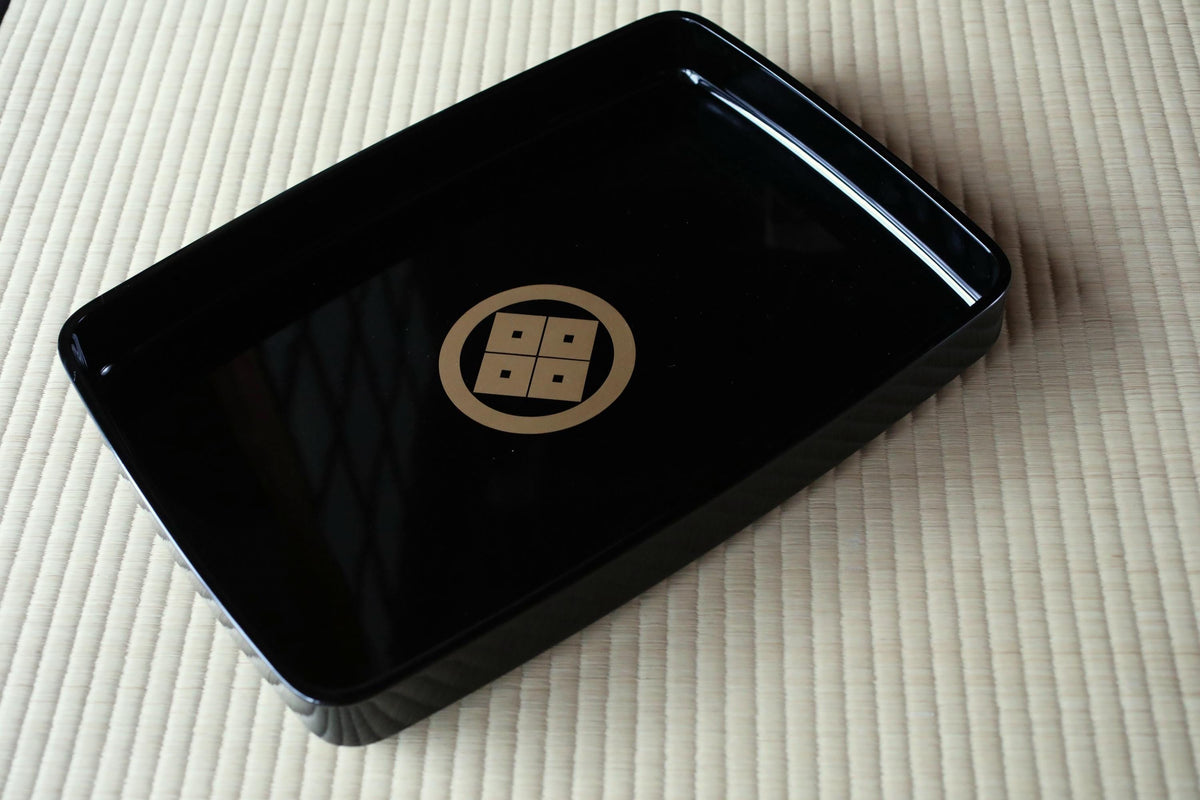 A black rectangular tray with a gold emblem on it, placed on a beige surface.