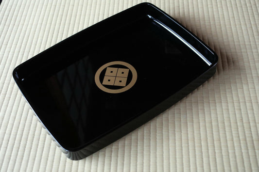 A black rectangular tray with a gold emblem on it, placed on a beige surface.
