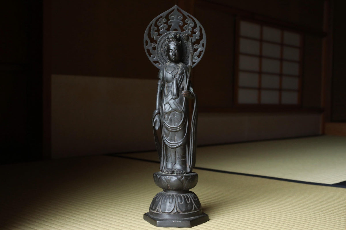 A black metal statue of a deity, possibly a Buddha, standing on a mat in a dimly lit room with a window in the background.