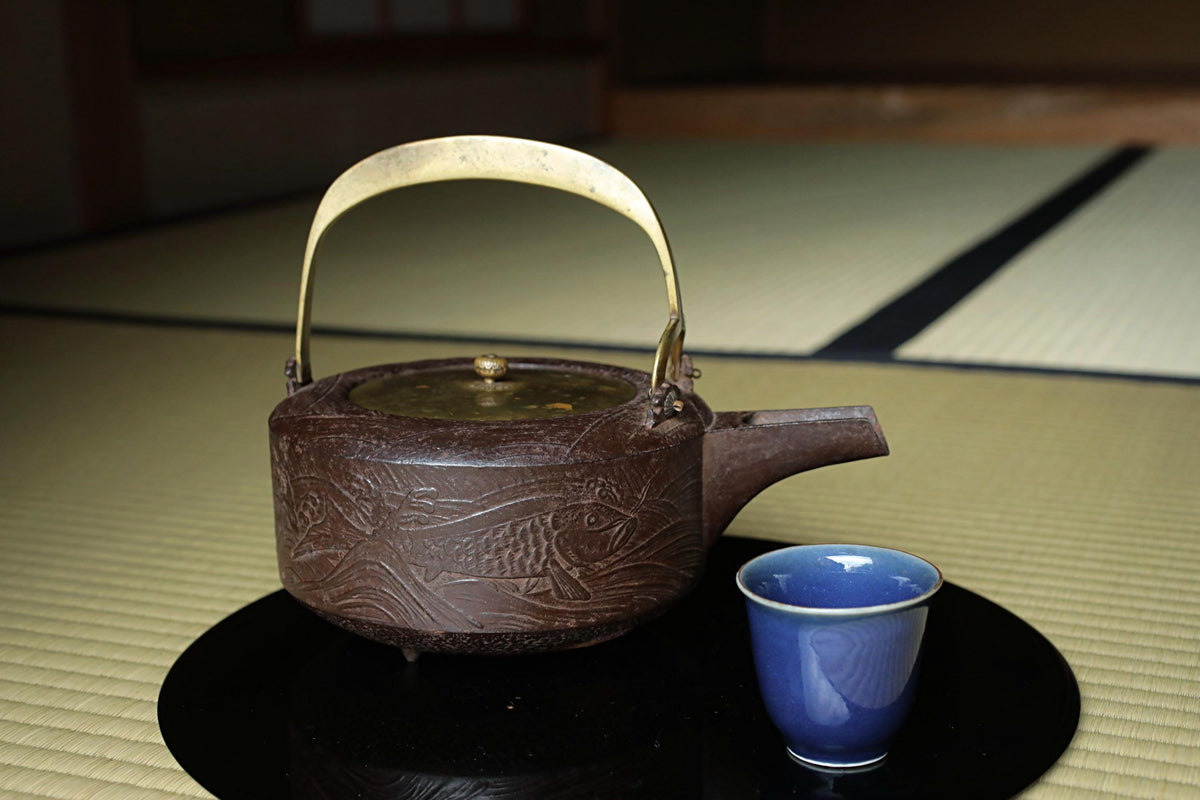 A traditional Japanese tea set consisting of a brown teapot with a gold handle and lid, and a blue ceramic cup, placed on a black tray on a tatami mat.