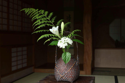 A traditional Japanese vase with a white flower and green ferns sits on a wooden surface, creating a serene and elegant atmosphere.
