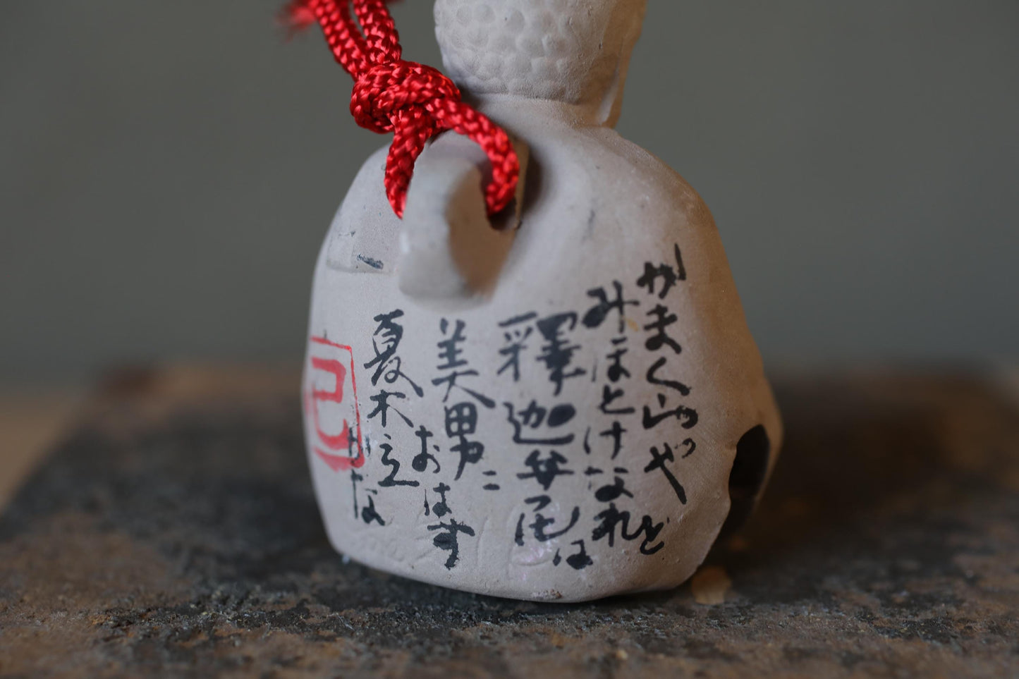 A close-up image of a ceramic bell with Japanese characters painted on it, and a red string tied around the bell.