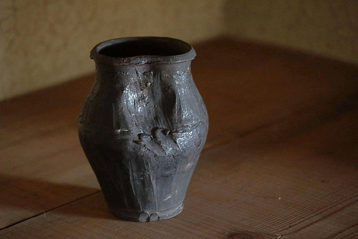 A dark-colored, weathered vase with a textured surface sits on a wooden surface.