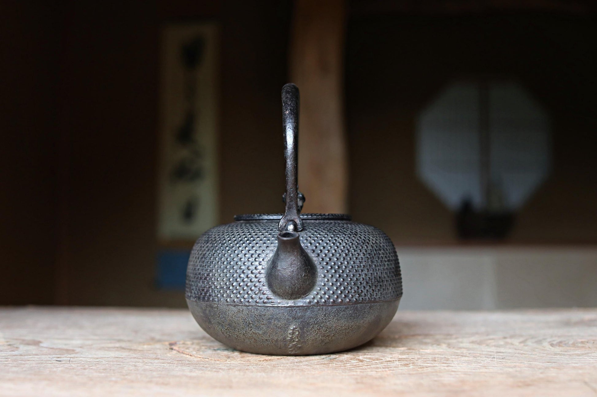 A black metal teapot with a textured surface sits on a wooden table, with a blurred background that includes a window and a wall.