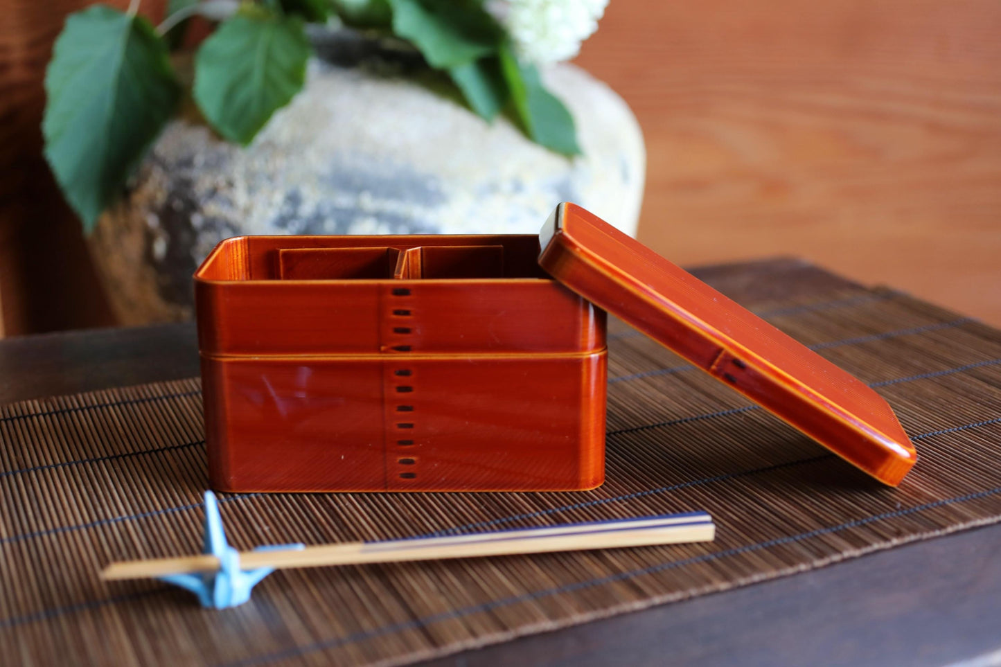 a wooden box with a lid, a pair of chopsticks, and a vase with green leaves on a table.