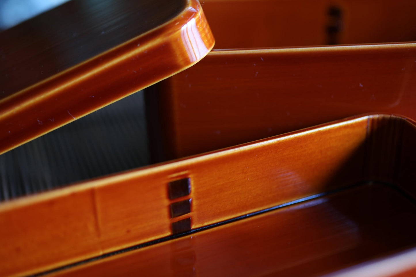 a close-up view of a wooden box with a shiny, orange-colored surface. The box appears to be made of wood and has a lid that is partially open, revealing the interior. The background is blurred, drawing focus to the box and its details.