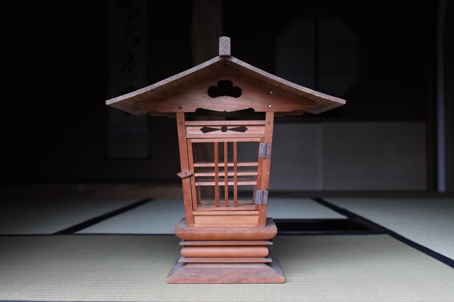 A wooden lantern with a pointed roof and intricate carvings sits on a tatami mat in a dimly lit room.