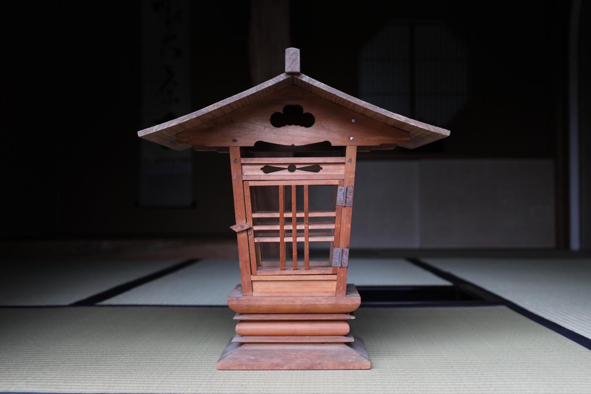 A wooden lantern with a pointed roof and intricate carvings sits on a tatami mat in a dimly lit room.