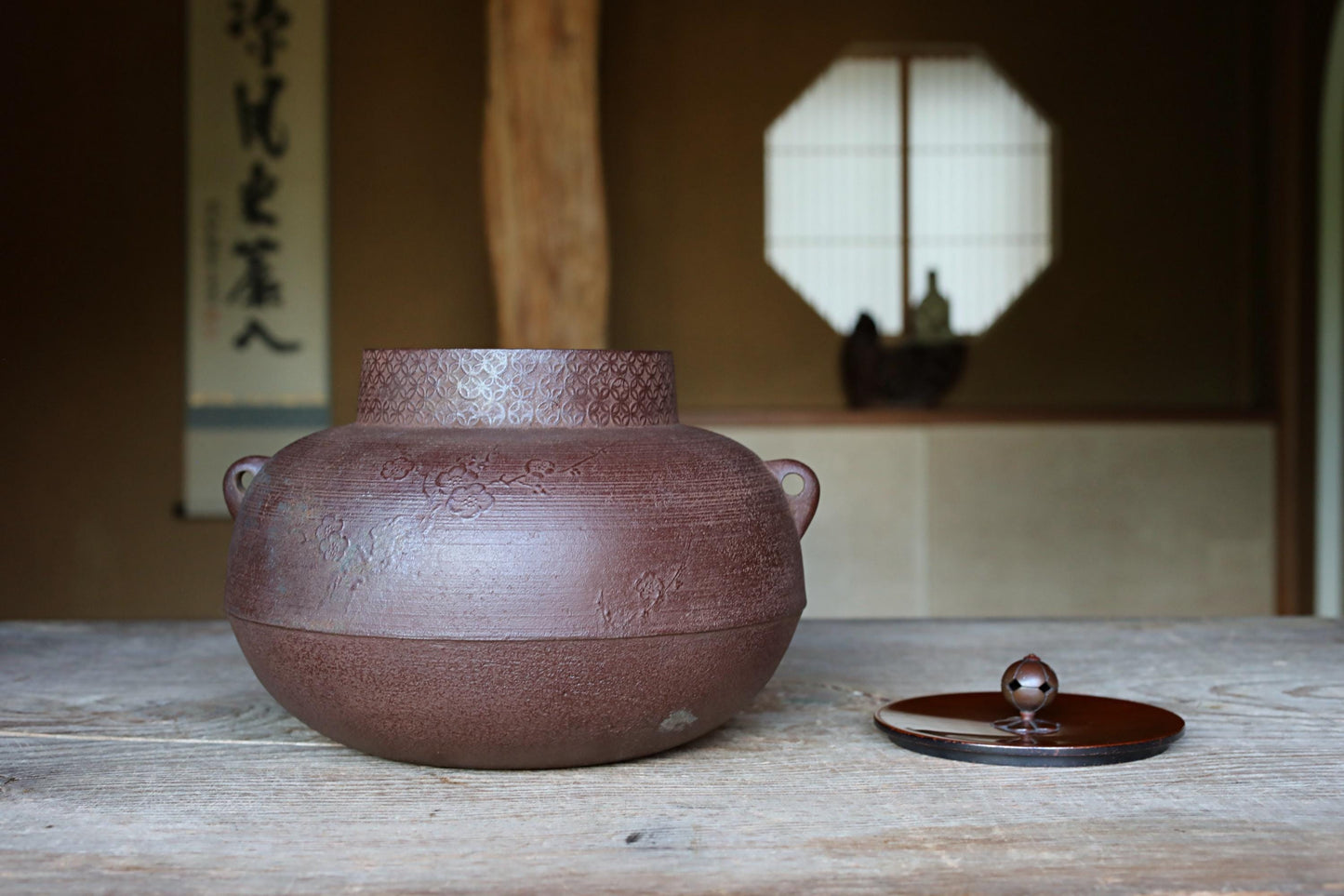 A brown ceramic tea pot with a lid sits on a wooden table, with a window and a wooden beam visible in the background.
