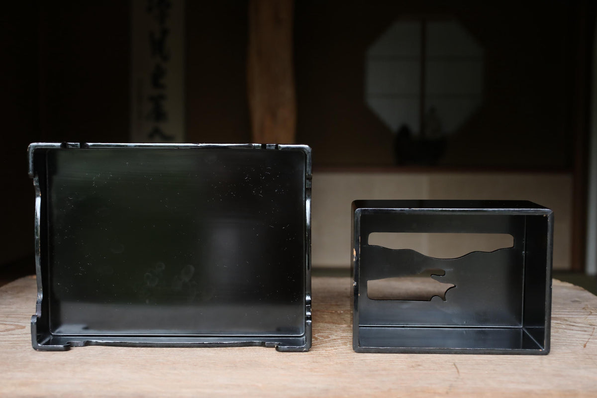 two black metal boxes on a wooden surface, one larger and one smaller, with a blurred background that appears to be a room with a window.