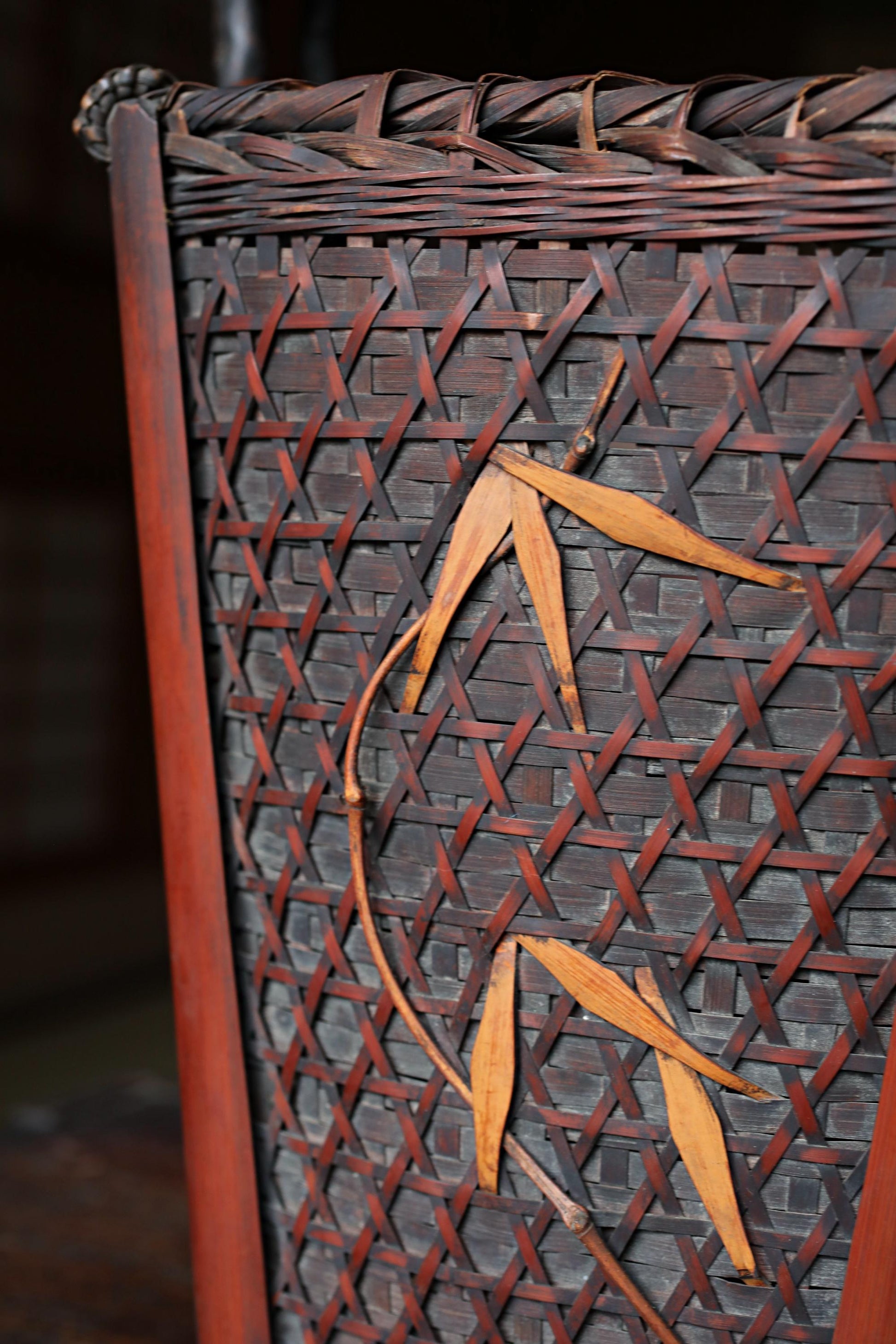 a close-up view of a woven basket or container with a distinctive pattern of brown and orange leaves or branches. The leaves are arranged in a repeating pattern, creating a visually striking design. The basket appears to be made of a material that resembles wood or bamboo, giving it a rustic and natural appearance.