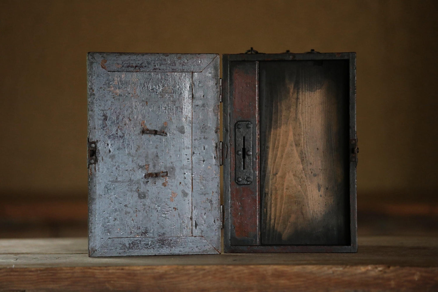 An old, weathered metal box with a wooden panel on the right side, sitting on a wooden surface against a plain background.