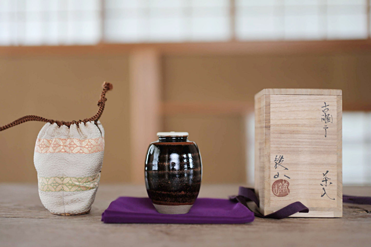 a wooden box with Japanese characters, a small ceramic jar, and a small bag or pouch on a purple cloth.