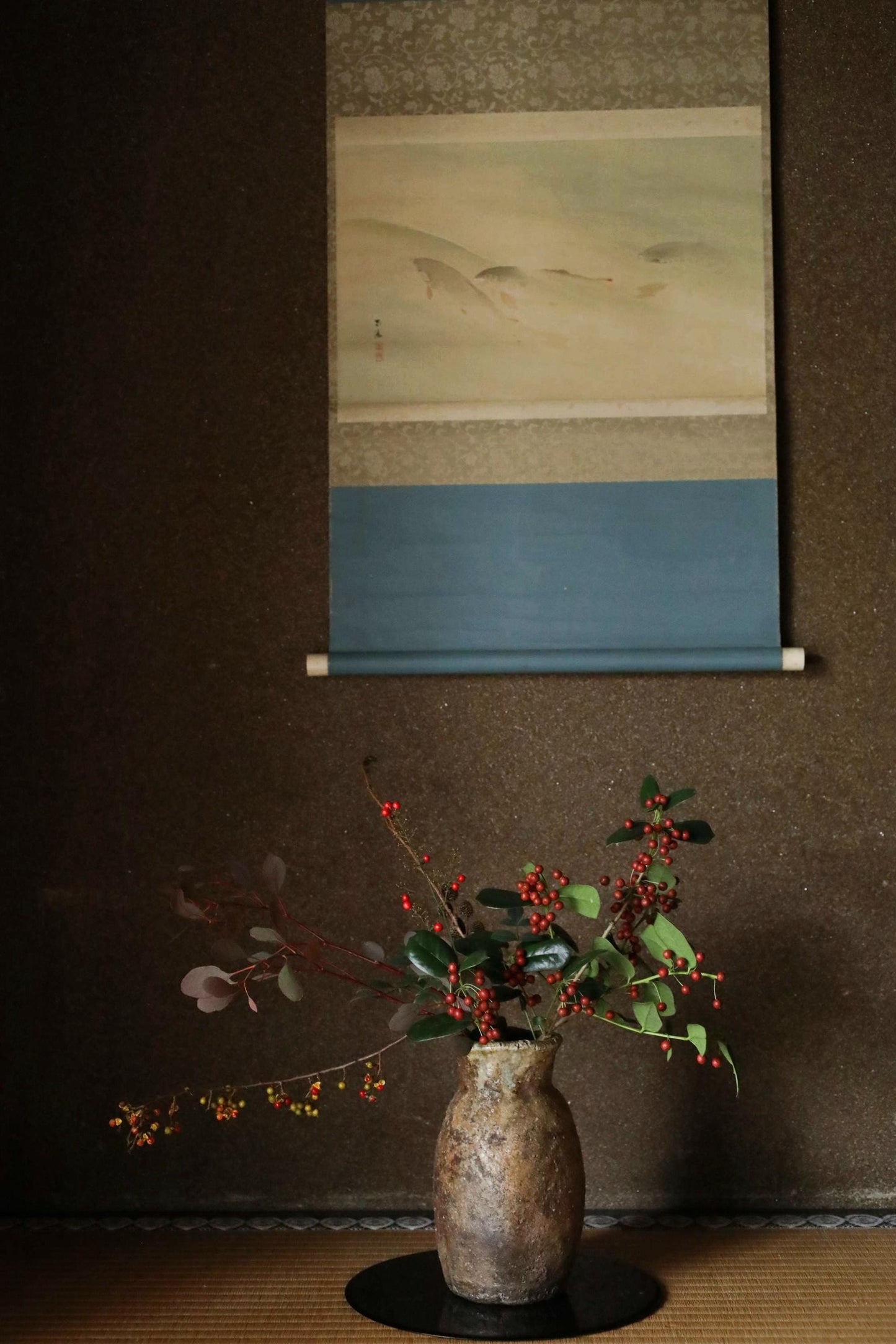 A vase filled with red berries and green leaves sits on a wooden surface, with a framed painting hanging above it.