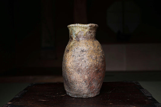 An old, weathered vase sits on a dark wooden surface, its surface showing signs of age and use.