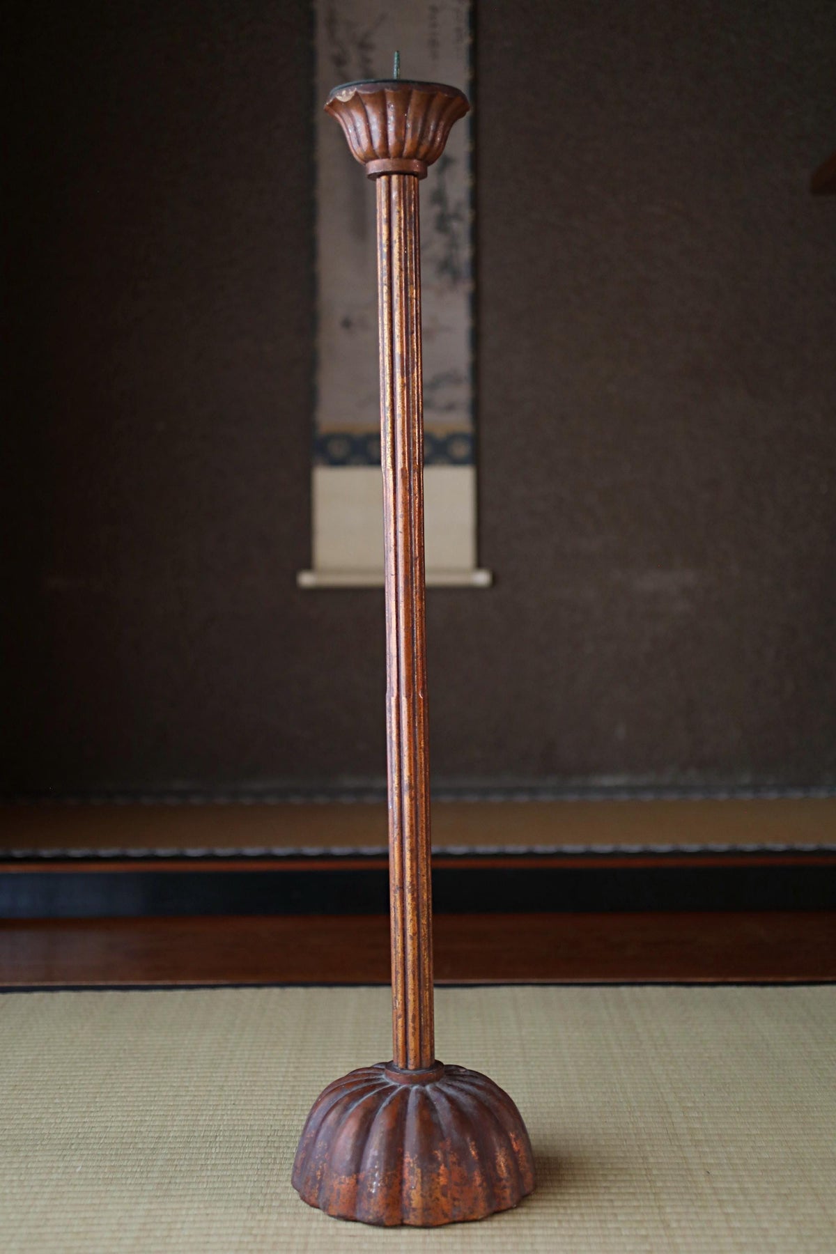 A tall, ornate wooden candle holder with a fluted design stands on a beige mat against a dark background.