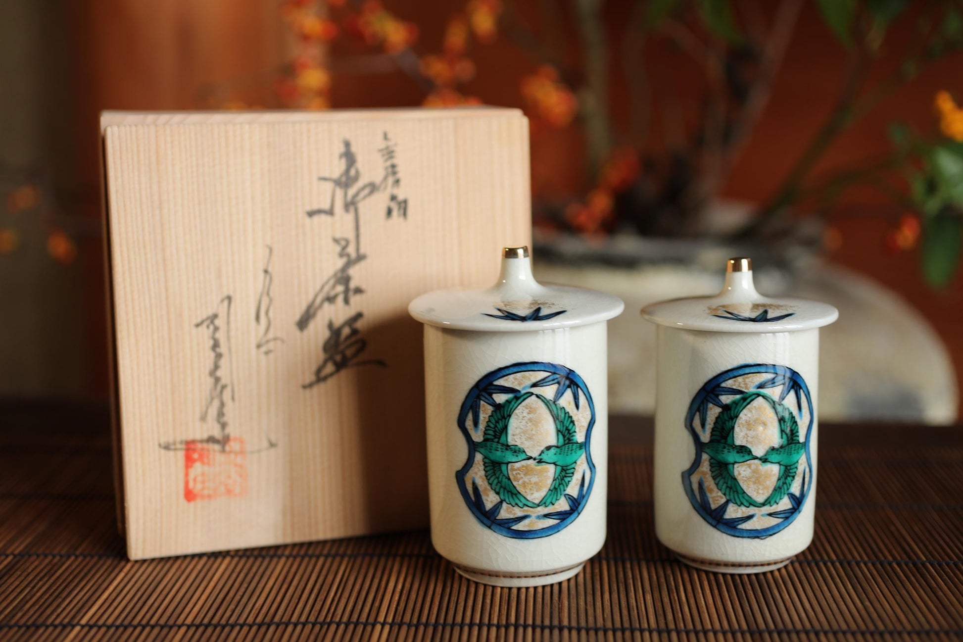 two white ceramic containers with blue and green designs, placed on a wooden surface. There is a wooden box with Japanese characters in the background.