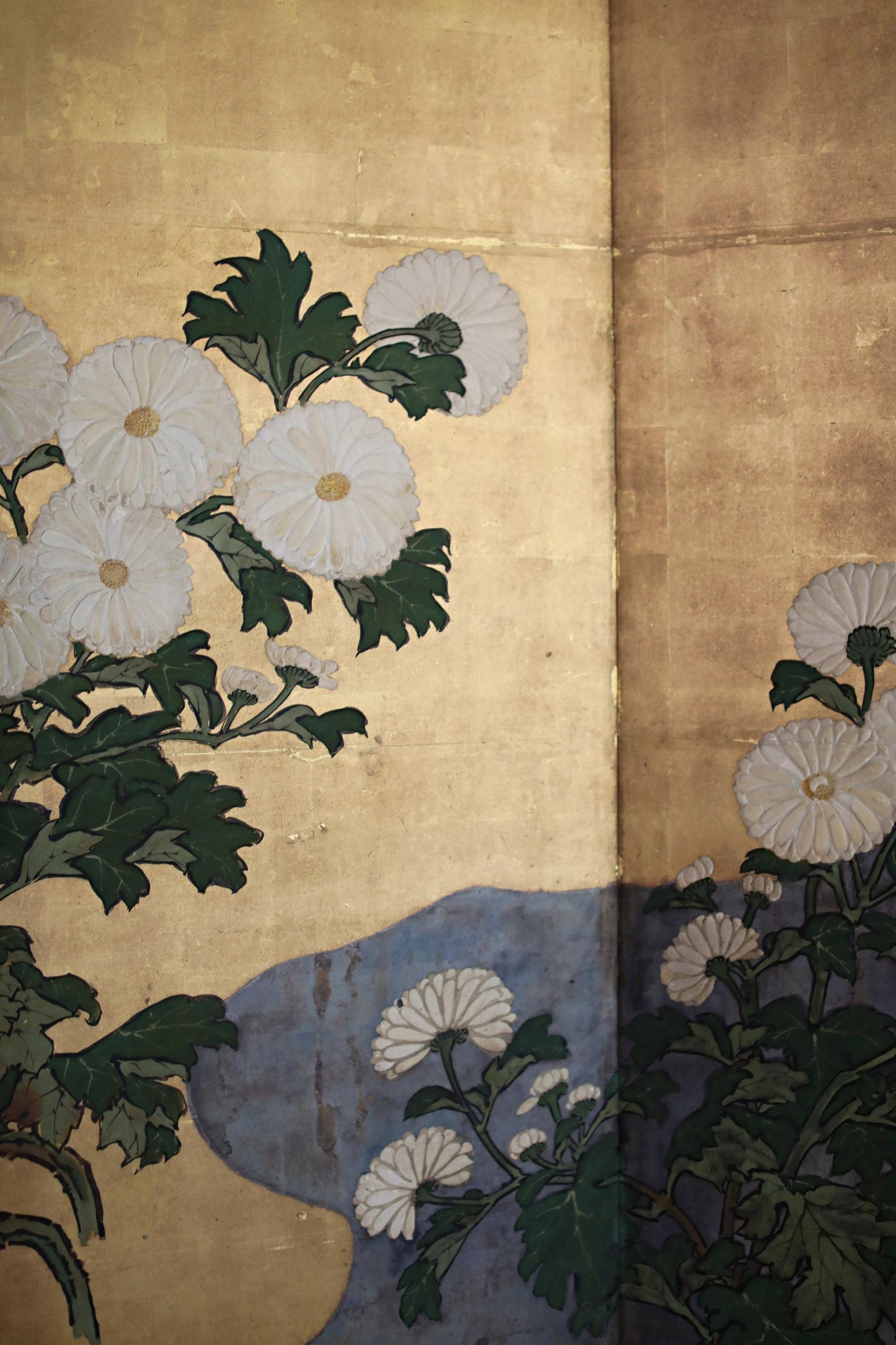 a close-up view of a painting or artwork depicting white flowers with green leaves. The flowers are arranged in a way that they appear to be growing out of a blue background, creating a visually appealing contrast. The overall composition of the image is simple and focused on the details of the flowers and the background.