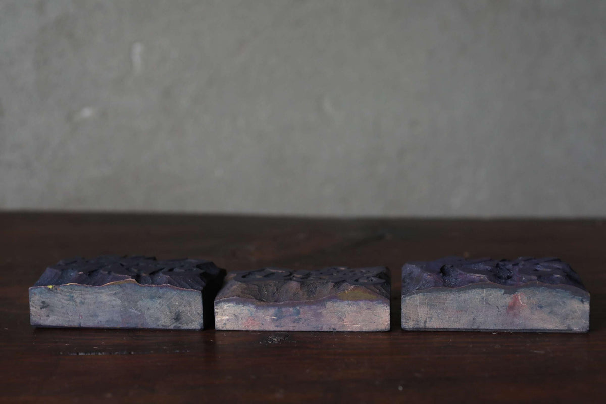 Three square metal blocks with a dark, textured surface are arranged on a wooden surface against a plain gray background.