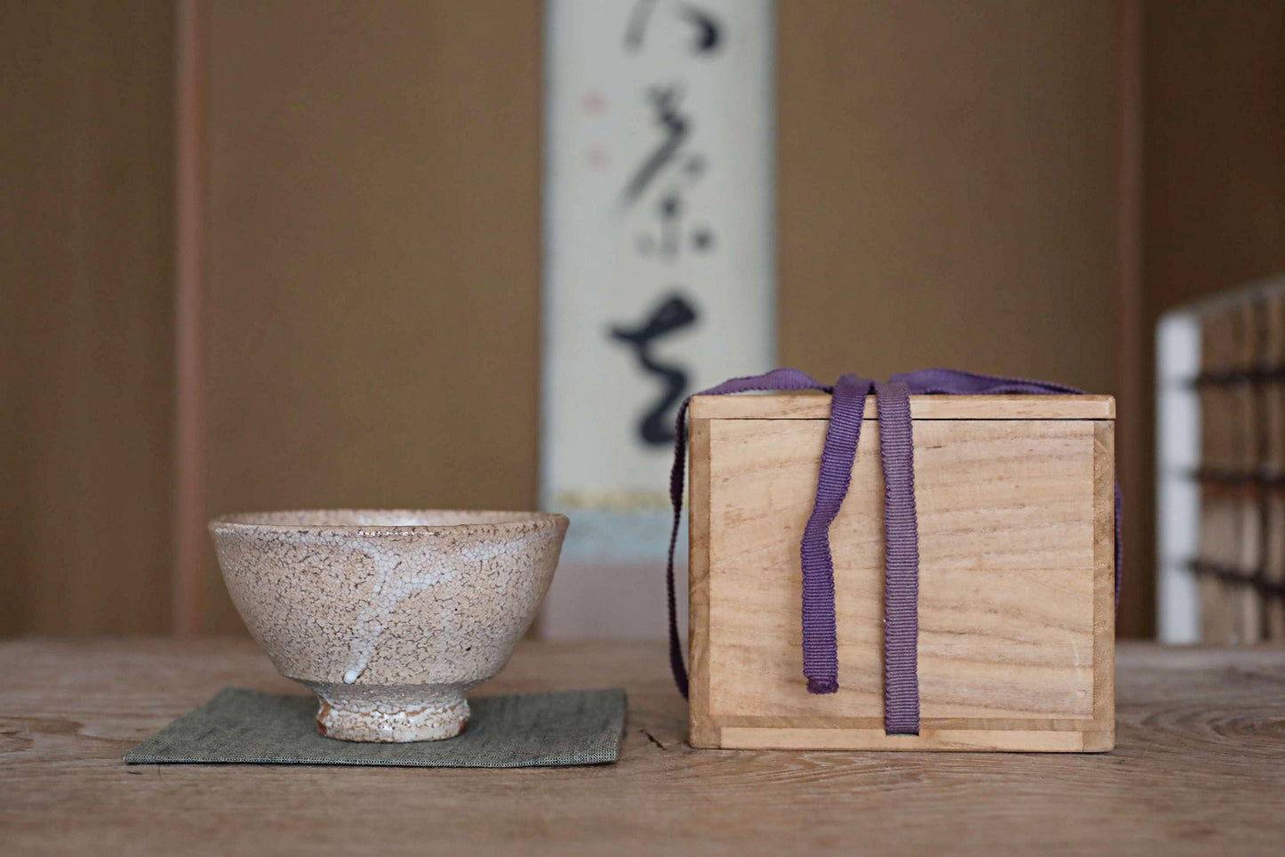 A white ceramic bowl and a wooden box with a purple ribbon are placed on a wooden table, with a white wall and a black and white Japanese calligraphy poster in the background.